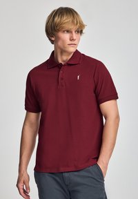 Polo de color burdeos hecho de algodón, con cuello, tapeta de tres botones y un pequeño logo blanco en el lado izquierdo del pecho. Mangas cortas.