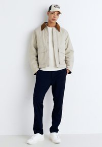 Chaqueta beige con cuello marrón, suéter blanco, pantalones azul marino y zapatillas blancas. Tejido texturizado con un corte casual, diseño minimalista.
