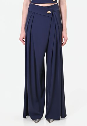 Pantalon large bleu marine avec détail noué et bouton doré à la taille, porté avec des chaussures métalliques à bouts pointus.