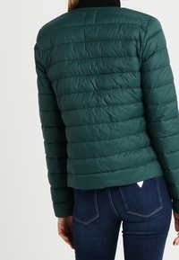 Doudoune verte avec un design matelassé. Elle présente un col montant et est associée à un jean en denim foncé, mettant en valeur une silhouette ajustée.
