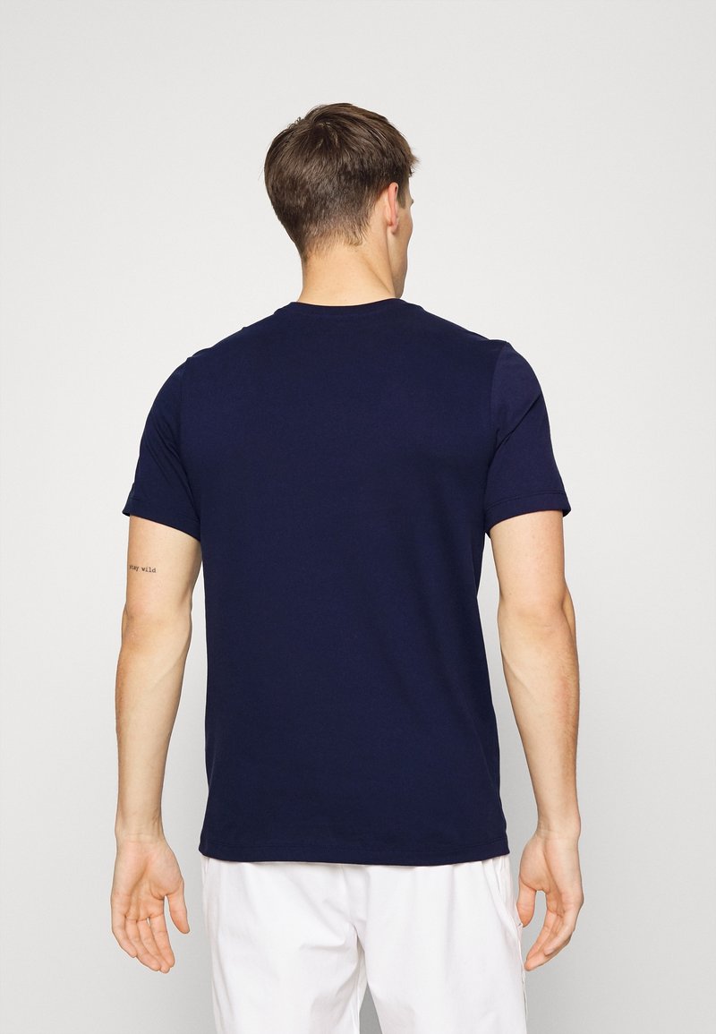 Homme aux cheveux courts et bruns portant un t-shirt bleu foncé et un pantalon blanc, debout de dos sur un fond uni.