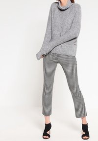 Pull gris à col drapé avec des poignets côtelés, associé à un pantalon noir et blanc à motif pied-de-poule longueur cheville et des escarpins noirs ouvertes.