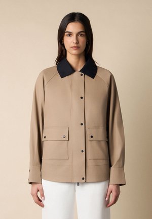 Kobieta zwrócona przodem, ubrana w beżową kurtkę z czarnym kołnierzem, czarnymi zatrzaskami, kieszeniami z patkami oraz białe spodnie na beżowym tle.