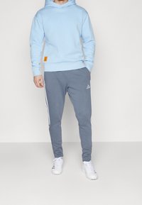 Felpa leggera blu chiaro con logo discreto, abbinata a pantaloni sportivi grigi con strisce laterali bianche e logo Adidas. Scarpe sportive bianche.