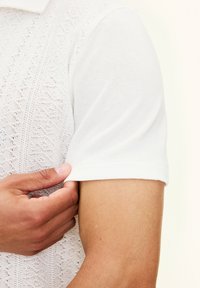 Biała, dzianinowa koszulka polo z krótkim rękawem, z wyraźnym wzorem. Kołnierzyk oraz krawędzie rękawów wykończone są gładko.