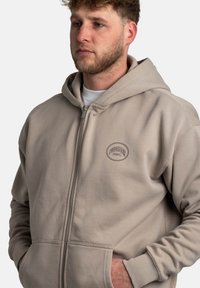 Sudadera cremosa con cremallera y capucha con cordón, que cuenta con un bolsillo frontal y un logotipo bordado de "Pescador AWG" en el pecho. Material de algodón suave.