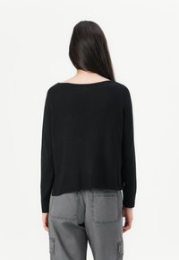 Femme avec de longs cheveux noirs portant un pullover noir à manches longues et un pantalon cargo gris, vue de dos sur un fond blanc.