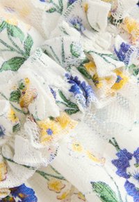 Weißer Stoff mit floralem Muster in Gelb und Blau, ausgestattet mit fransigen Kanten und einer strukturierten, leicht durchsichtigen Oberfläche.