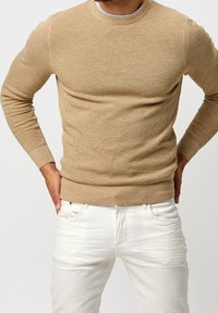 Beige stickad tröja med rundad halsringning och ribbad fåll, tillsammans med vita jeans. Tröjan har ett strukturerat, lätt material.