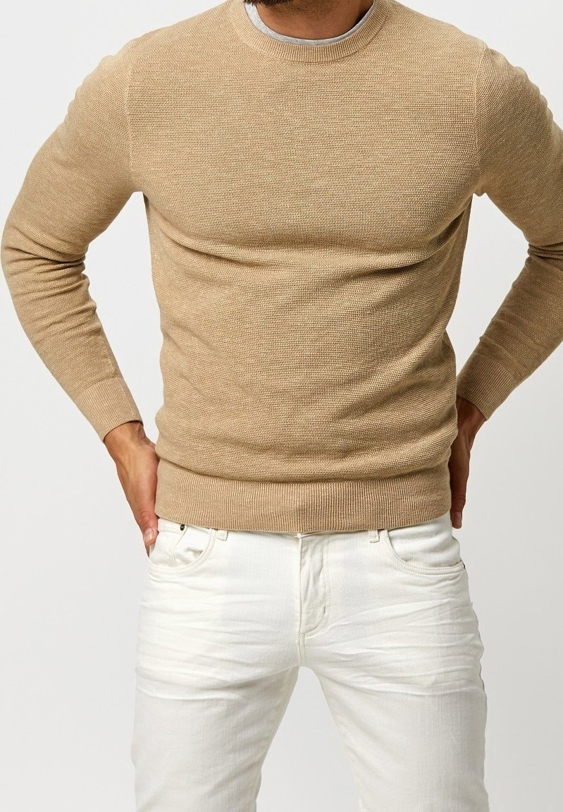 Beige stickad tröja med rundad halsringning och ribbad fåll, tillsammans med vita jeans. Tröjan har ett strukturerat, lätt material.