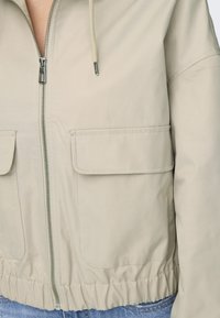Beige bunda se zipovým zapínáním vpředu, stahovací kapucí a dvěma velkými klopovými kapsami. Má elastický spodní lem a hladkou texturu látky.