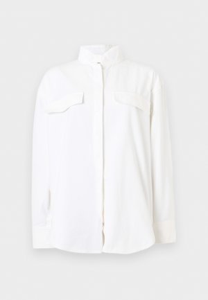 Camicia bianca a maniche lunghe con bottoni, due tasche sul petto e collo alto, mostrata su uno sfondo bianco neutro.
