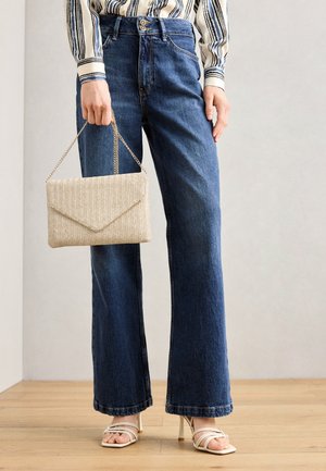 Kvinde iført blå jeans med vide ben, hvide høje sandaler med stropper, stribet skjorte, som holder en beige struktureret håndtaske med kæderem.