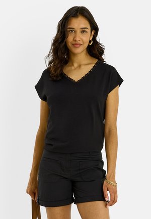 Mujer con cabello oscuro ondulado, vistiendo una camiseta negra de manga corta con escote en V y pantalones cortos negros, de pie frente a un fondo claro y liso.