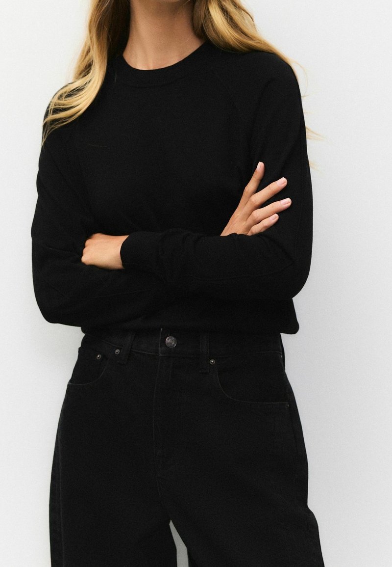 Personne avec de longs cheveux blonds portant un pull noir à manches longues et un pantalon noir taille haute, les bras croisés devant un fond blanc.