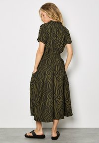 Olivengrünes Midikleid mit schwarzen gewellten Streifen, kurzen Ärmeln, Gummibund und seitlichen Taschen, kombiniert mit schwarzen Rutschen-Sandalen.