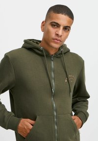 Felpa verde con zip, dotata di tasca a marsupio, cappuccio regolabile con cordino e dettaglio del logo sul petto. Realizzata in tessuto morbido e testurizzato.