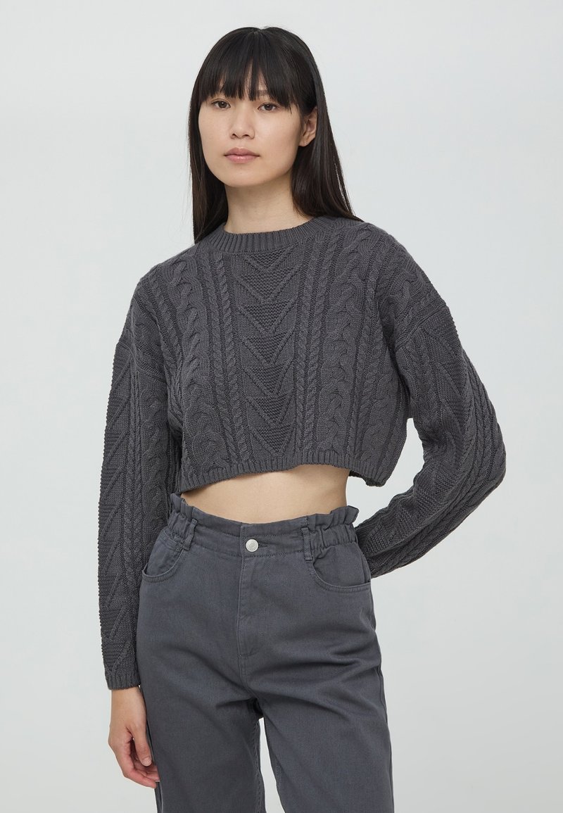 Maglione grigio corto con design a trecce testurizzate, scollatura rotonda e maniche lunghe, abbinato a pantaloni grigi a vita alta con cintura arricciata.