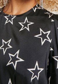 Svart tygtröja med ett stjärnmönster i vitt, med en slät yta och rund halsringning. Detaljer inkluderar plisseringar vid axlarna.