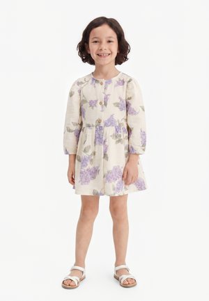 Fată tânără zâmbitoare cu păr scurt și închis la culoare, purtând o rochie crem cu imprimeu floral mov și sandale albe, stând în fața unui fundal alb.