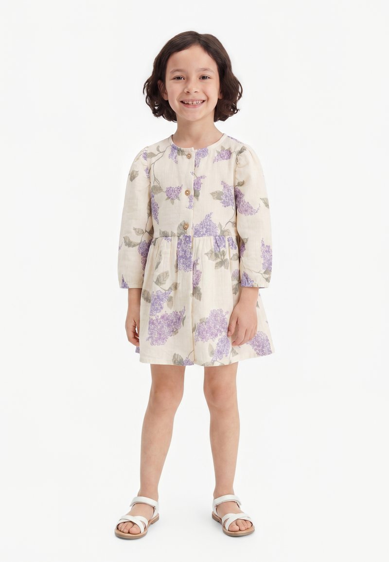 Jeune fille souriante aux cheveux courts et foncés, portant une robe crème à imprimé fleuri violet et des sandales blanches, debout devant un fond blanc.