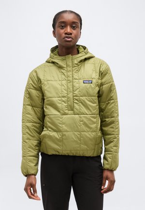 Žena v olivovo zelenej prešívanej kapucňovej bunde Patagonia a čiernych nohaviciach stojaca pred jednoduchým svetlosivým pozadím.