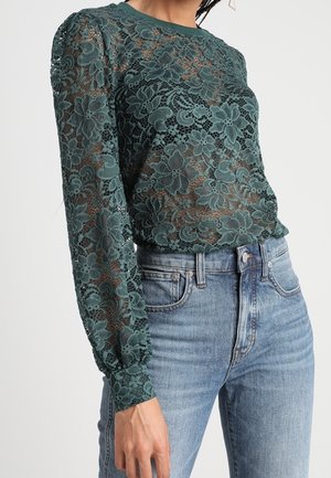 Blouse en dentelle fleurie vert foncé à manches longues bouffantes, associée à un jean en denim bleu clair taille haute.