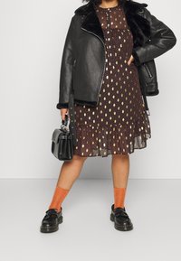 Giubbotto di pelle nera con colletto in pelliccia sintetica, vestito marrone con pois dorati, calze arancioni, scarpe nere e una piccola borsa nera.