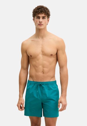 Jeune homme aux cheveux bouclés portant un short de bain turquoise, debout devant un fond uni, face à l'avant avec les bras détendus.