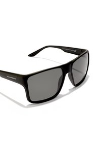 Ochelari de soare din plastic negru cu lentile întunecate, ramă pătrată și brațe texturate, cu marca "HAWKERS" pe lateral.