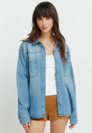 Junge Frau mit langem braunem Haar, die ein hellblaues Jeanshemd über einem weißen Top und braune Shorts trägt, vor einem schlichten Hintergrund stehend.