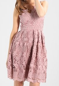 Höyhenenvaaleanpunainen pitsimekko, jossa on kukkakuviot, hihaton malli, siron aaltoileva helma ja vartalonmyötäinen yläosa levenevällä helmalla.