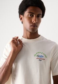 Krämlik bomull t-shirt med färgglada broderade texter och grafik som står "Scotch & Soda Amsterdam 1985." Rund hals, kort ärm.