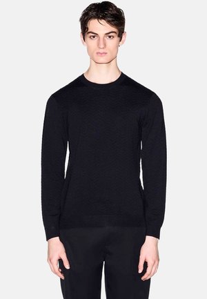 Junger Mann trägt einen enganliegenden schwarzen, strukturierten Langarmpullover und schwarze Hose, steht vor einem schlichten hellen Hintergrund.