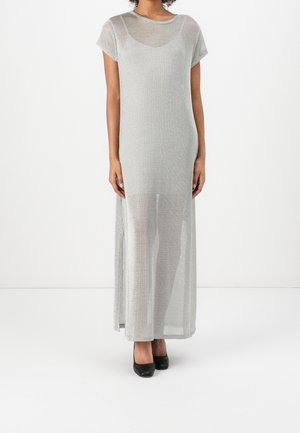 Femme portant une robe longue argentée à manches courtes et transparente par-dessus une combinaison noire, assortie à des talons fermés noirs, debout sur un fond blanc.