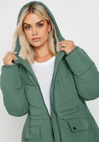 Grön puffjacka med huva, med dragkedja framtill, sidfickor och en quiltad textur. Bärs över en vit skjorta.