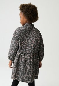 Dijete s kovrčavom kosom okrenuto u stranu, nosi haljinu dugih rukava s uzorkom sive i crne leopardske dlake s elastičnim manžetnama.