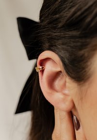 Goldene Creolen mit drei klaren Edelsteinen, die am Ohrläppchen getragen werden. Der Hintergrund zeigt dunkles Haar und eine schwarze Schleifenbinde.