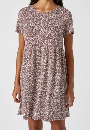 Robe à manches courtes au-dessus du genou avec un petit imprimé floral blanc sur fond couleur rouille portée par une femme debout.