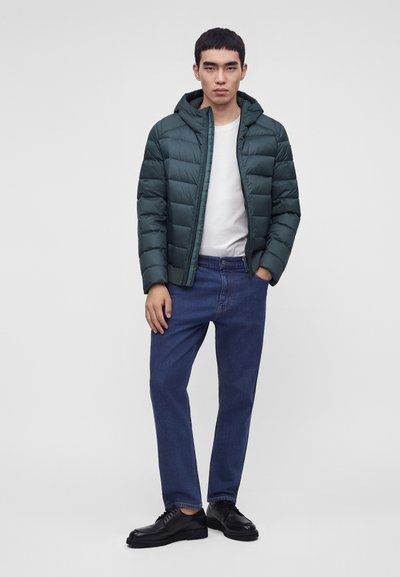 Homme portant une doudoune vert foncé sur une chemise blanche, un jean bleu et des chaussures noires, debout devant un fond clair uni.
