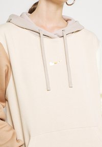 Beige hoodie med ljusbruna accenter. Har en dragsko i huvan och en liten gyllene Nike-logotyp på bröstet. Mjukt, texturerat tyg.