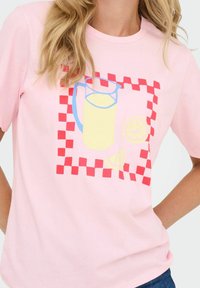 Lichtroze t-shirt met een grafisch ontwerp van een kan en citrusplakken binnen een rood en roze geruit kader. Korte mouwen.