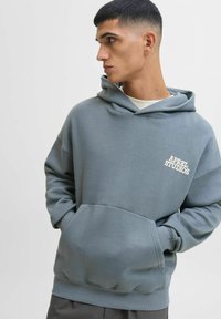 Šedá mikina s kapucí a přední kapsou, s bílým logem "APREL STUDIOS". Měkká směs bavlny, volný střih, pružný lem a manžety.