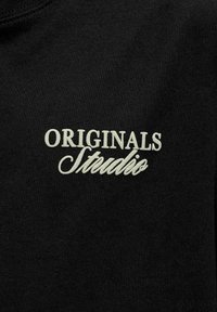 Tela negra con texto blanco "ORIGINALS Studio" impreso en dos fuentes diferentes, mayúsculas y escritura cursiva.