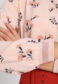 Blusa rosa chiaro con motivo floreale in blu scuro e arancione, caratterizzata da maniche arricciate e polsini lavorati a maglia con un accento glitterato.