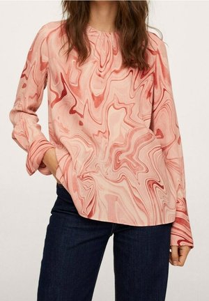 Woman wearing a long-sleeve, pink and red swirl-patterned blouse and dark blue jeans with one hand in pocket against plain background.