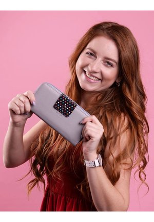 Gray wallet with a colorful polka dot accent on the clasp. Features a zipper closure and is held by a person against a pink background.