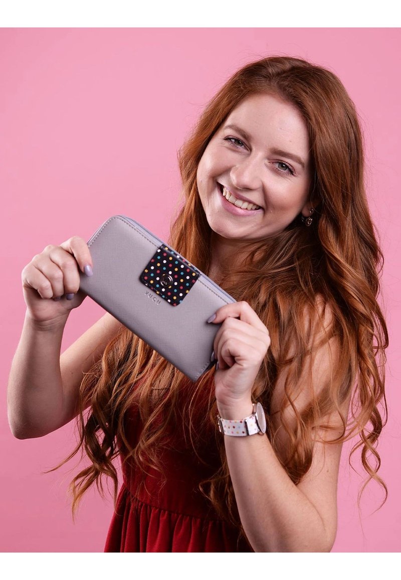 Gray wallet with a colorful polka dot accent on the clasp. Features a zipper closure and is held by a person against a pink background.