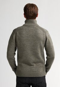 Bărbat văzut din spate purtând un pulover tricotat verde măslin cu guler înalt și pantaloni negri, pe un fundal deschis simplu.