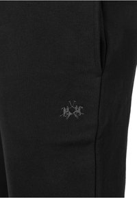 Svarta mjukisbyxor med texturerat tyg, med en liten grå logotyp av två ryttare på hästar på vänstra låret nära fickan.